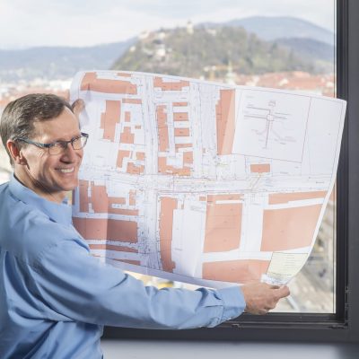Mitarbeiter, der einen Stadtplan in den Händen hält und vor einem Fenster mit Blick auf den Grazer Schlossberg steht