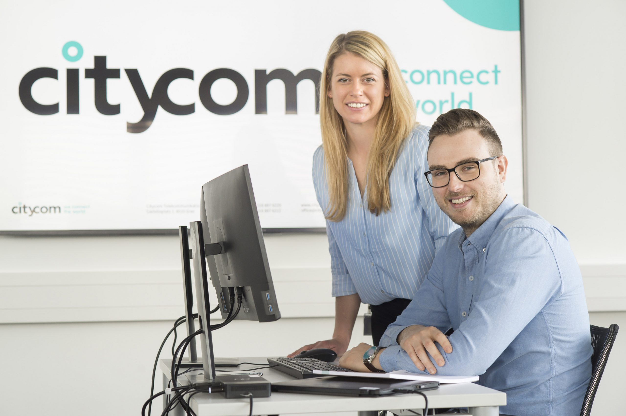 Citycom Mitarbeiterin steht neben ihrem Kollegin am Schreibtisch und beide lachen in die Kamera