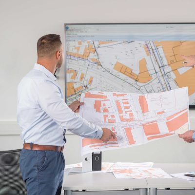 Zwei Citycom Mitarbeiter, die einen Stadtplan halten und auf einem großen Bildschirm einen anderen Plan betrachten